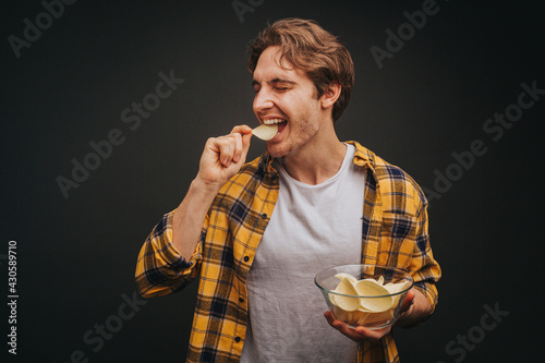 Young blond man in yellow shirt is eating chips and holding plate with it, isolated over black background