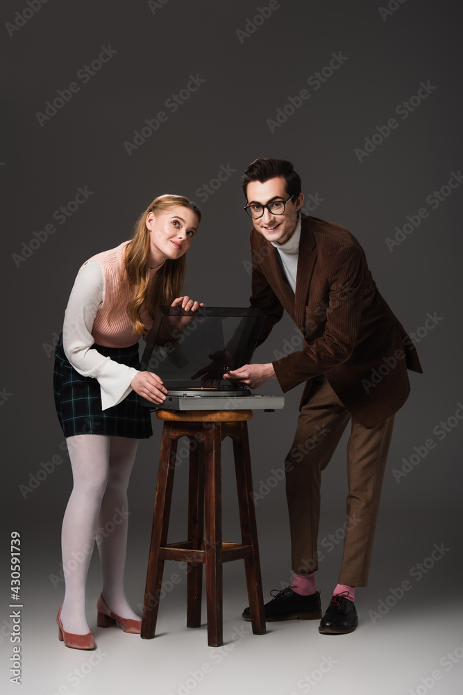 Obraz premium full length view of happy couple in retro outfit near record player on dark grey background