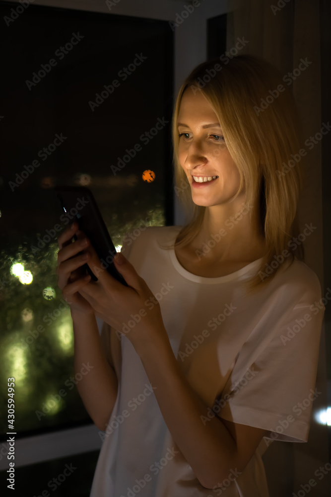 Portrait of a beautiful young blonde woman with an open smile in a white shirt with a mobile phone in her hands near the window of a night city background, warm light from the phone falls on her face Portrait of a beautiful young blonde woman with an open smile in a white shirt with a mobile phone in her hands near the window of a night city background, warm light from the phone falls on her face