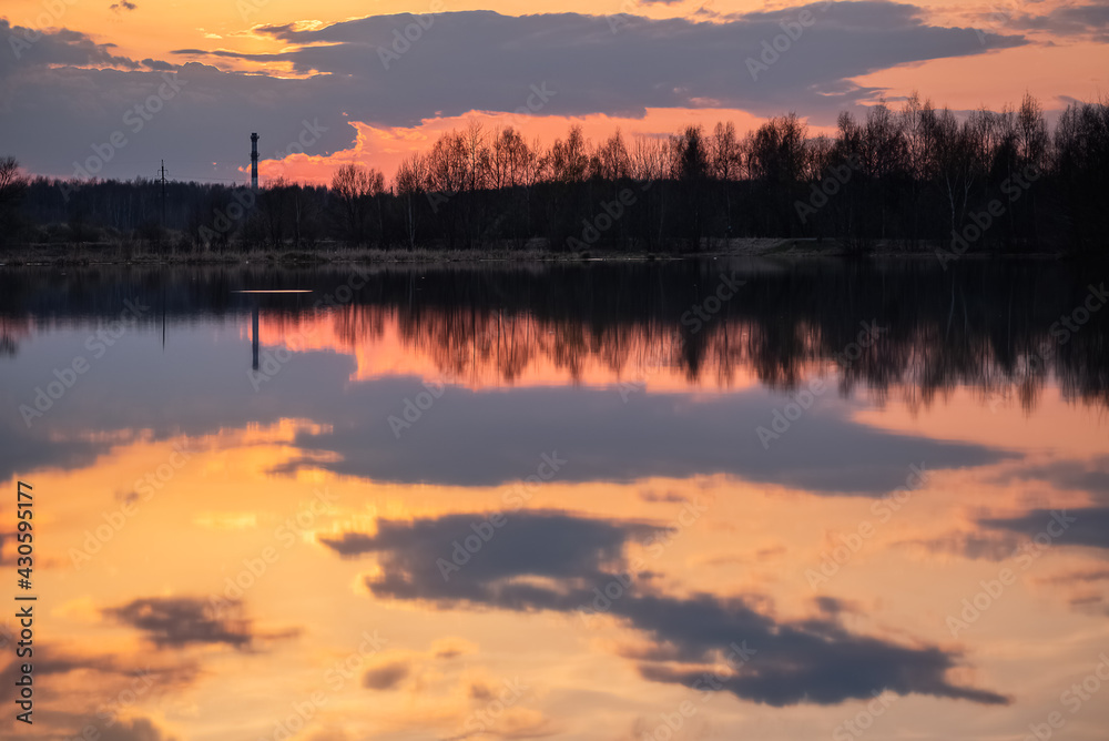 Dramatic sunset sky, clouds reflect in still water surface of forest lake