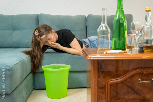 Dark-haired young woman is lying on the sofa and she is ill from alcohol intoxication, she is going to vomit over a bucket, there are empty bottles in the background