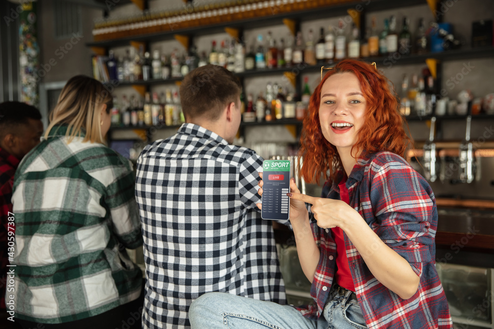 Woman showing phone with betting app on screen. Emotional fans cheering for favourite team, watching on exciting game. Concept of friendship, leisure activity, emotions Woman showing phone with betting app on screen. Emotional fans cheering for favourite team, watching on exciting game. Concept of friendship, leisure activity, emotions