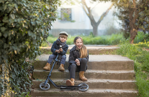 a boy and a girl sit on the steps with a scooter. they make faces and have fun. They're wearing vintage clothes.