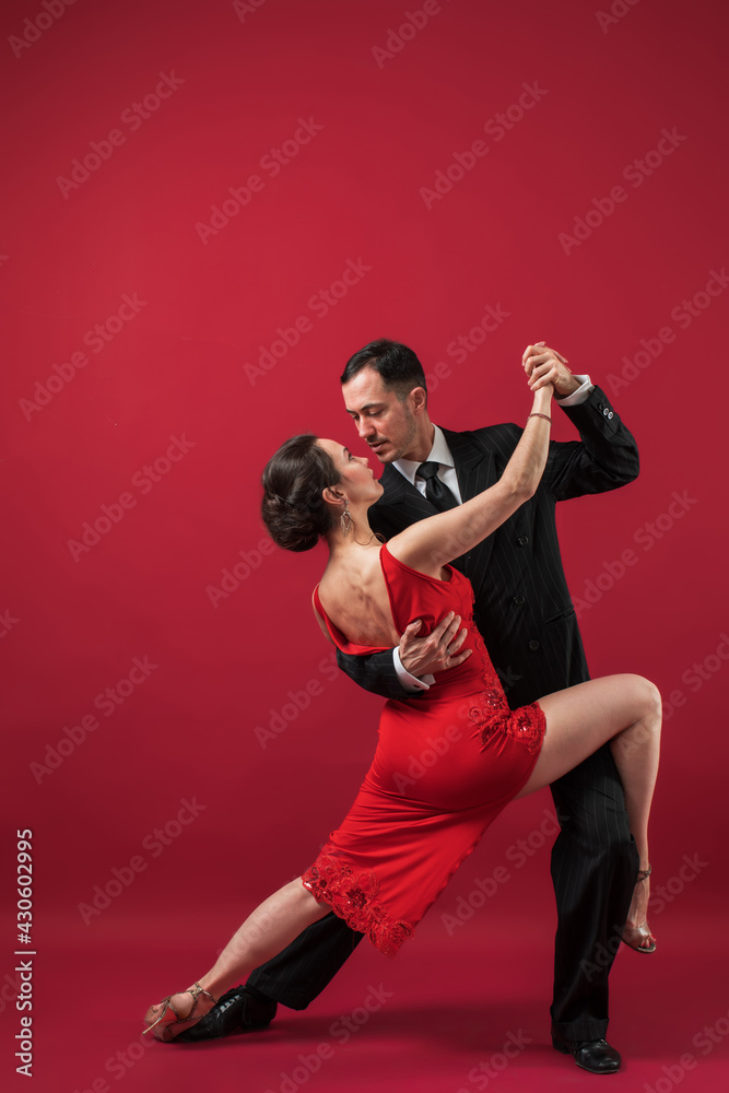 Couple of professional tango dancers in elegant suit and dress pose in a dancing movement on red background. Handsome man and woman dance looking  eye to eye.