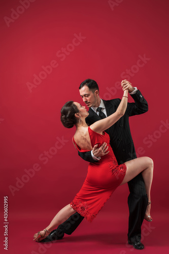 Couple of professional tango dancers in elegant suit and dress pose in a dancing movement on red background. Handsome man and woman dance looking eye to eye.
