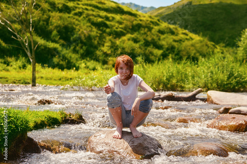 Fresh water drops. Blurred young  girl with red hair sits on a stone in the river and splash water drops. Concept of summer rest on the beach  good weather  happiness. Altay Republic  Siberia