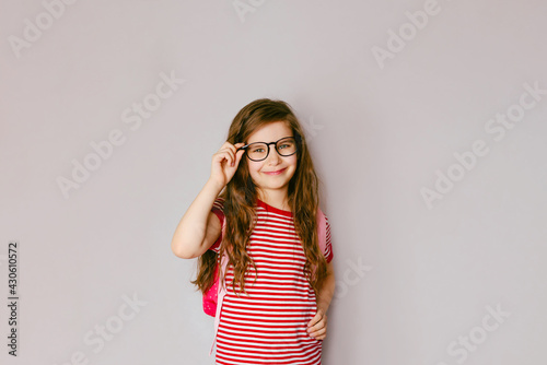 Back to school. Portrait of a blonde schoolgirl 6-7 y.o. in black glasses with a bag with books. Grey Studio background. Education. Smiling at the camera. Copyspace.