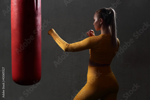 Young beautiful sports girl in a yellow tight-fitting body tracksuit beats a red punching bag in the gym without gloves. Fitness trainer shows exercises. European girl works out in the fitness center.