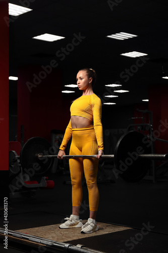 Fototapeta Naklejka Na Ścianę i Meble -  Young beautiful sportive girl in a yellow tight-fitting body tracksuit does deadlift in the gym. Fitness trainer shows exercises. European girl works out in the fitness center. Pulls the barbell