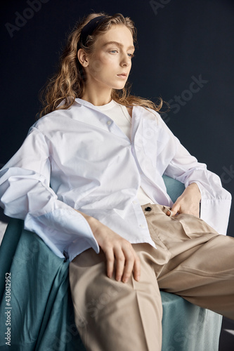 portrait shot in the studio on a dark background of a beautiful young blonde girl with big blue eyes, long wavy hair, she is wearing a white cotton shirt with a T-shirt under it and beige pants