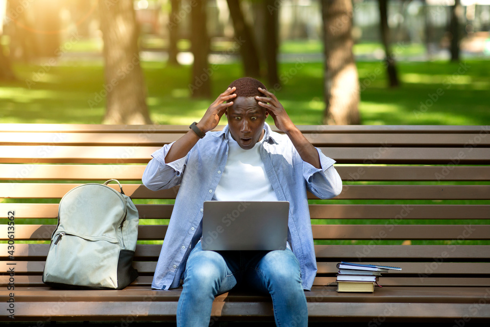 Desperate black guy with laptop holding his head in panic on bench at park