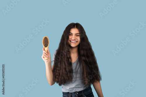 Portrsit of beautiful Indian teen girl with long dark hair holding wooden brush and smiling at camera on blue background photo