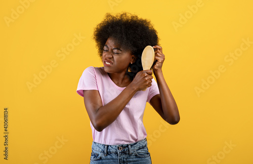 Annoyed african american woman having problem brushing her bushy curly hair on yellow studio background