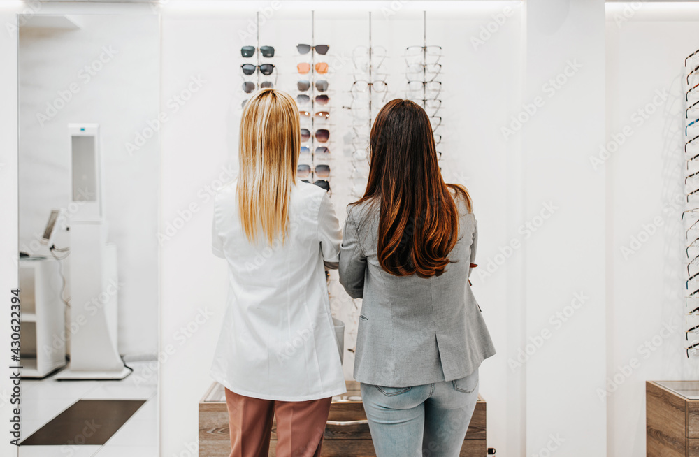 Fototapeta premium Beautiful and fashionable woman choosing eyeglasses frame in modern optical store. Female seller specialist helps her to make right decision.