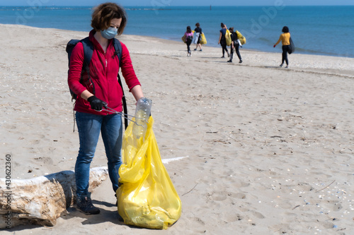 Donna che pulisce la spiaggia mettendo una bottiglia di plastica nel sacco dei rifiuti, a Lido di Dante in Italia, e sullo sfondo ci sono altre persone che portano sacchi di rifiuti. Senza plastica.  photo