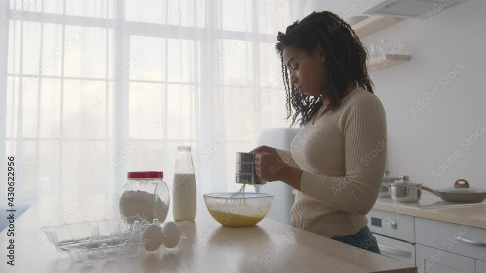 Young concentrated african american woman cooking at kitchen, mixing dough in bowl and adding some flour with sifter