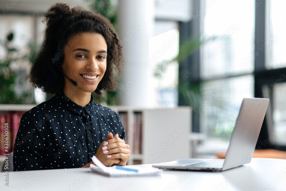 Portrait of attractive young african american woman in headset, manager, call center worker or freelancer, sitting in office, wearing stylish clothes, using laptop, looking at camera, smiling