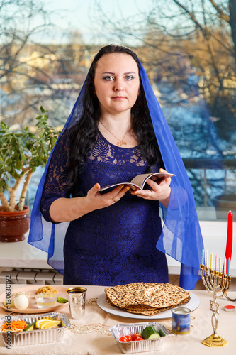 A Jewish woman with her head covered in a blue cape at the Passover Seder table reads the Passover Haggadah.