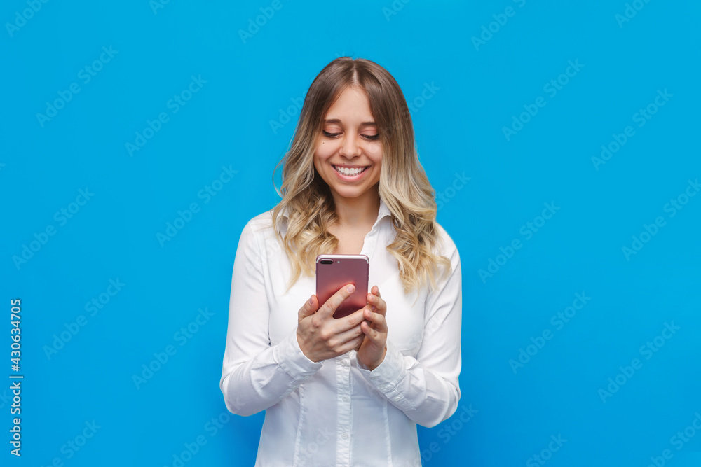 A young caucasian happy pretty blonde woman in a office white shirt smiles and holds a mobile phone looking at the screen isolated on a bright color blue background. Student girl uses her smartphone A young caucasian happy pretty blonde woman in a office white shirt smiles and holds a mobile phone looking at the screen isolated on a bright color blue background. Student girl uses her smartphone