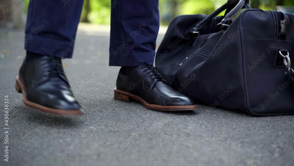 Close-up of male feet in leather shoes standing outdoors with travel bag. Unrecognizable confident elegant businessman on business trip on sunny summer day. Business lifestyle Close-up of male feet in leather shoes standing outdoors with travel bag. Unrecognizable confident elegant businessman on business trip on sunny summer day. Business lifestyle