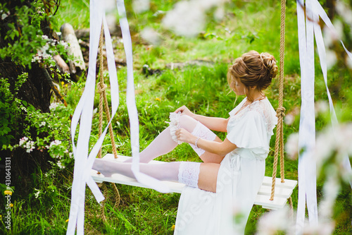Young bride with blond hair in white negligee and stockings putting on a garter