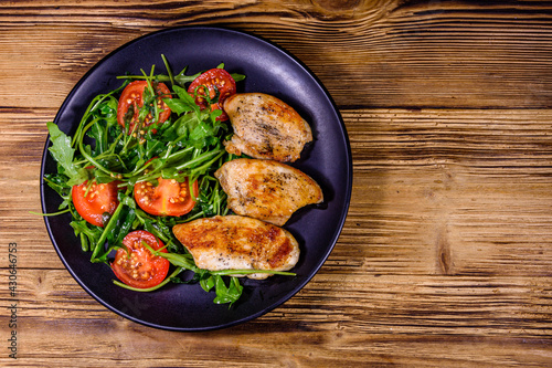 Roasted chicken breasts and salad with arugula and cherry tomatoes in a black plate. Top view