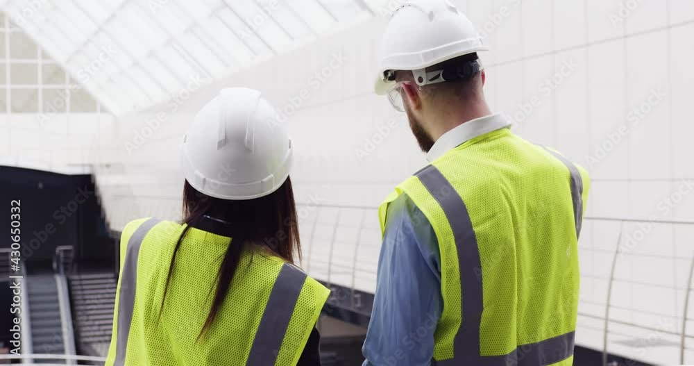 Rear view of male civil engineer and female architect wearing hard hats standing inside new office center construction site, discussing project design details. Rear view of male civil engineer and female architect wearing hard hats standing inside new office center construction site, discussing project design details.