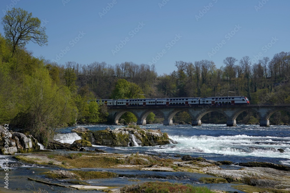 Neuhausen, Switzerland - 04 24 2021: Rhine river near Rhine Falls. There is Rhine bridge at town Laufen constructed for rail transport and pedestrians. Swiss federal railways train passes through. Neuhausen, Switzerland - 04 24 2021: Rhine river near Rhine Falls. There is Rhine bridge at town Laufen constructed for rail transport and pedestrians. Swiss federal railways train passes through.