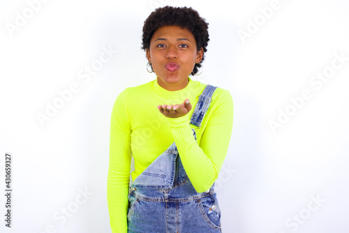 young African American woman with short hair wearing denim overall against white wall looking at the camera blowing a kiss with hand on air being lovely and sexy. Love expression.