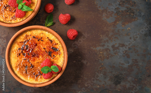 Fotografie Creme brulee desserts with cream and cane sugar with fresh raspberries and mint leaves in clay bowls on a dark vintage table, mock up with copy space