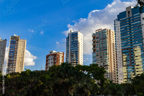 Detalhes de pr  dios entre   rvores com c  u azul e algumas nuvens ao fundo.