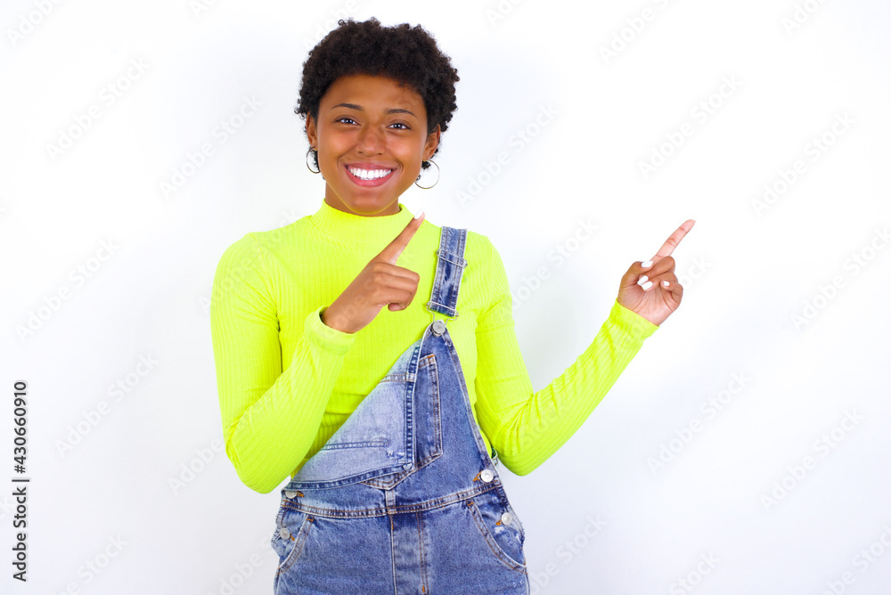 Positive young African American woman with short hair wearing denim overall against white wall with beaming smile pointing with two fingers and looking on empty copy space. Advertisement concept. Positive young African American woman with short hair wearing denim overall against white wall with beaming smile pointing with two fingers and looking on empty copy space. Advertisement concept.