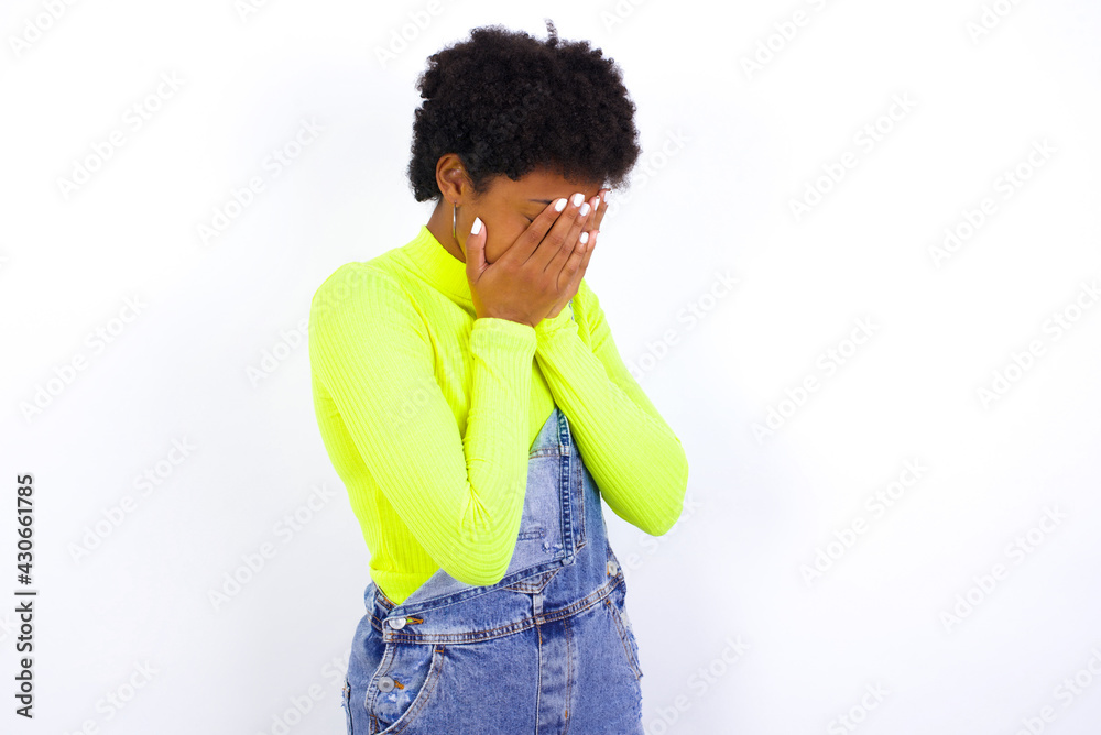 Sad young African American woman with short hair wearing denim overall against white wall covering face with hands and crying.