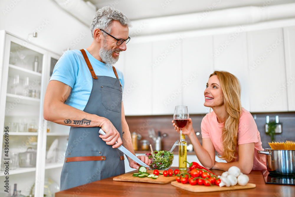 Happy senior couple in the kitchen. Mature man in apron preparing romantic dinner for his wife. Vegan, vegetarian, healthy lifestyle concept. Happy senior couple in the kitchen. Mature man in apron preparing romantic dinner for his wife. Vegan, vegetarian, healthy lifestyle concept.