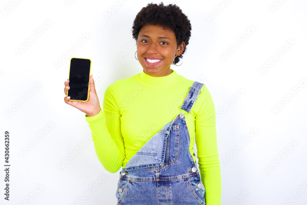 Smiling young African American woman with short hair wearing denim overall against white wall showing empty phone screen. Advertisement and communication concept. Smiling young African American woman with short hair wearing denim overall against white wall showing empty phone screen. Advertisement and communication concept.