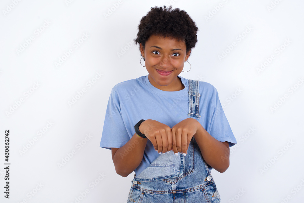 African American female with afro hair wears jeans overalls over white wall makes bunny paws and looks with innocent expression plays with her little kid African American female with afro hair wears jeans overalls over white wall makes bunny paws and looks with innocent expression plays with her little kid