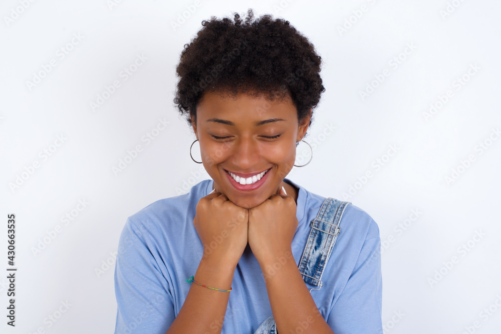 Cheerful young African American woman with short hair wearing denim overall against white wall has shy satisfied expression, smiles broadly, shows white teeth, People emotions Cheerful young African American woman with short hair wearing denim overall against white wall has shy satisfied expression, smiles broadly, shows white teeth, People emotions