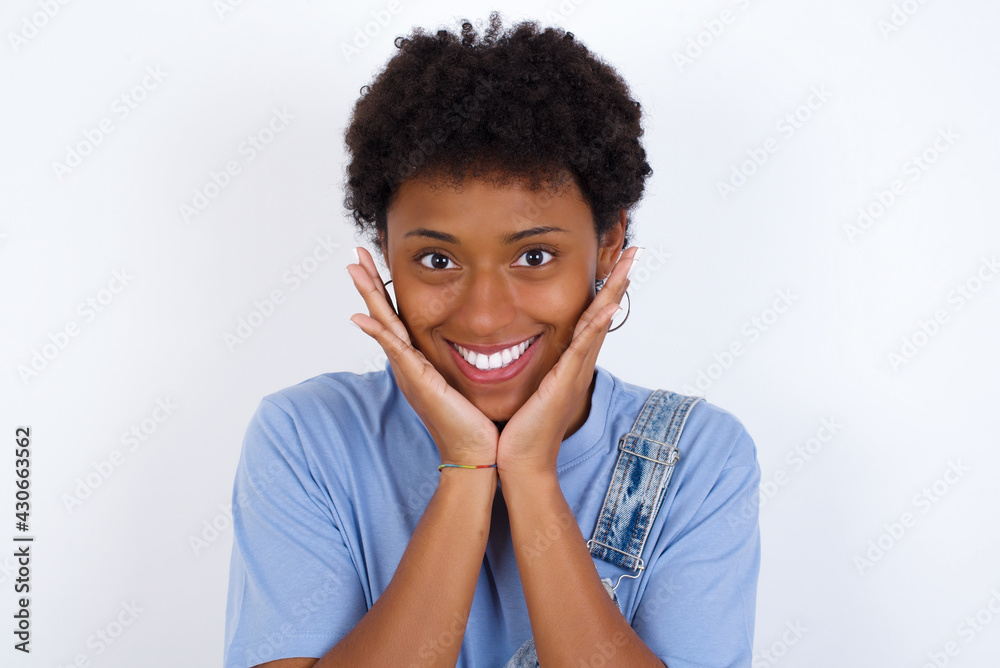Happy young African American woman with short hair wearing denim overall against white wall touches both cheeks gently, has tender smile, shows white teeth, gazes positively straightly at camera, Happy young African American woman with short hair wearing denim overall against white wall touches both cheeks gently, has tender smile, shows white teeth, gazes positively straightly at camera,