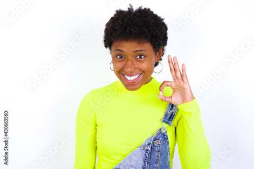African American female with curly bushy wears jeans overalls over white wall hold hand arm okey symbol toothy approve advising novelty news