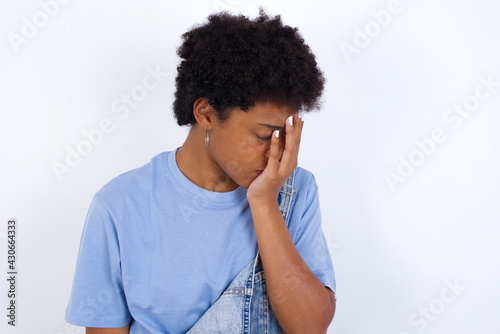 young African American woman with short hair wearing denim overall against white wall with sad expression covering face with hands while crying. Depression concept.