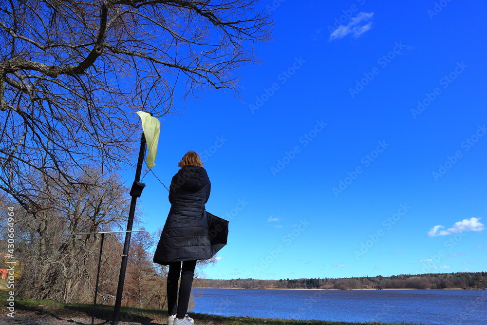 Swedish girl looking at the landscape. Sunny spring weather outside. Trees and a small hill. Dark gray jacket with a hood. Lake or water in distance. Skokloster, Håbo, Uppsala, Sweden, Europe.