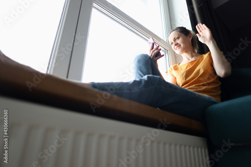 Young woman sitting on windowsill and waving her hand at screen of mobile phone