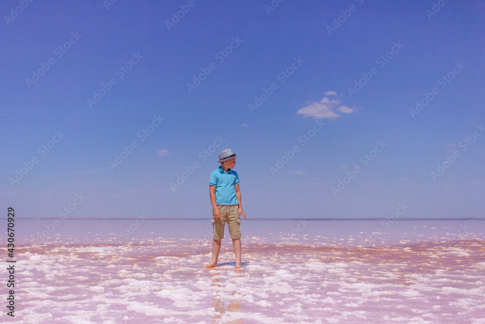 happy teenager in blue T-shirt, beige shorts and hat standing on an amazing pink lake with sea water and blue sky