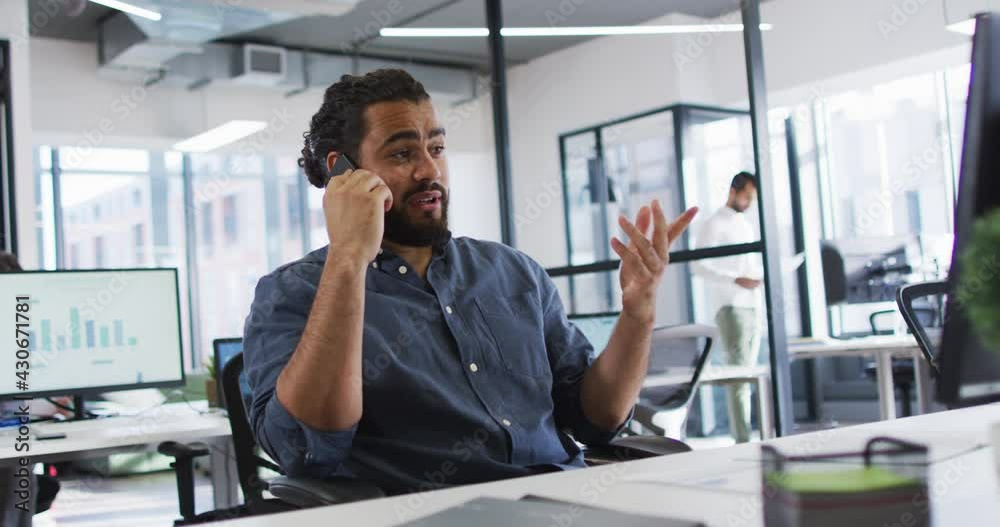 Mixed race businessman sitting at desk looking at computer screen, using smart phone and smiling Mixed race businessman sitting at desk looking at computer screen, using smart phone and smiling