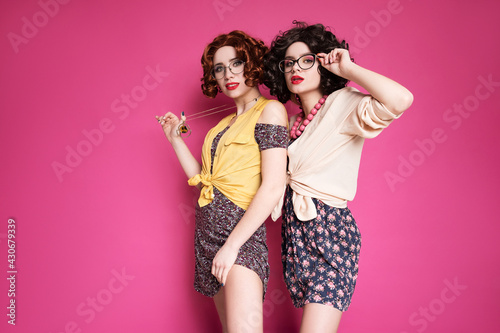 Two cute girl friends woman looking like nerd accountants standing on a pink background. They wear curly brunette wigs and unstylish retro casual outfits.