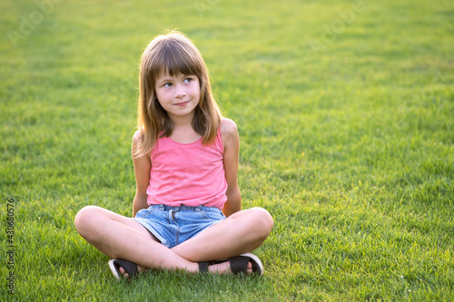 Young happy child girl resting while sitting on green grass lawn on warm summer day. © bilanol