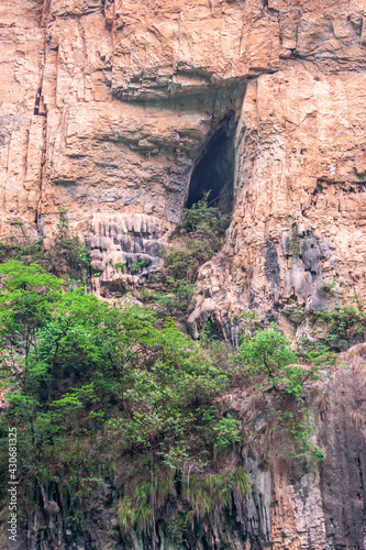 Wushan, China - May 7, 2010: Dawu or Misty Gorge on Daning River. Portrait of black cave hole in redish rock cliff with green foliage at bottom.