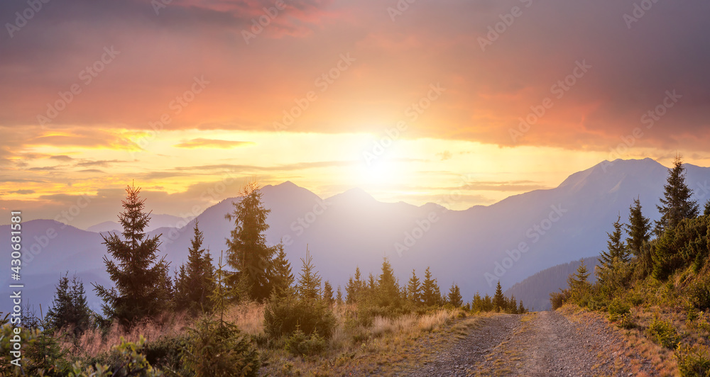 Panorama of warm sunsetover high mountain hills with a gravel road in front.