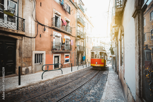 A wide-angle view of a red retro tram on a narrow street with one-way rail traffic in a European city; a vintage tourist streetcar in red and yellow colors on a tramway track over paving stone, Lisbon