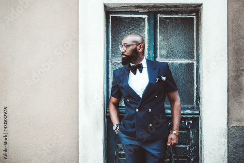A handsome dapper bald black guy with a beard, in an elegant dark-blue suit with a bow-tie and glasses, has just left a house through a peeled antique door and looking aside